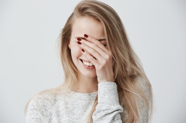 Gut aussehende emotionale junge kaukasische Frau mit blonden langen Haaren, die Augen mit der Hand schließend, während sie laut lacht, glücklich mit guten positiven Nachrichten, breit lächelnd, gerade weiße Zähne zeigend