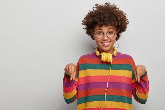 Gut aussehende ethnische weibliche Punkte zeigen mit den Zeigefingern nach unten, teilen ein schönes Angebot, freuen sich, tragen eine Brille und einen Pullover, isoliert über weißem Hintergrund.