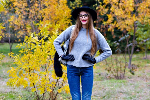 Gut aussehende Frau mit langen Haaren trägt Jeans und Handschuhe, die in sicherer Haltung auf Naturhintergrund stehen. Foto im Freien des hübschen weiblichen Modells im trendigen grauen Pullover, der im Park am Herbsttag geht.
