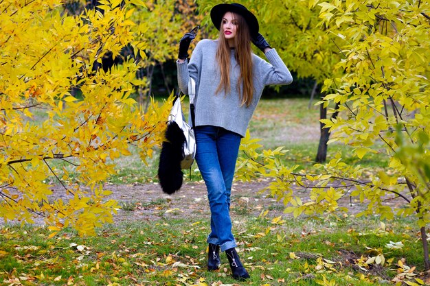 Gut aussehende Frau mit langen Haaren trägt Jeans und Handschuhe, die in sicherer Haltung auf Naturhintergrund stehen. Foto im Freien des hübschen weiblichen Modells im trendigen grauen Pullover, der im Park am Herbsttag geht.