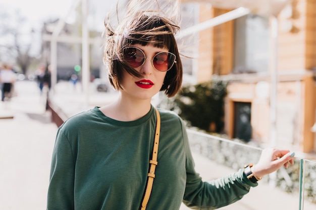 Gut aussehende junge Frau in der Sonnenbrille, die mit Interesse schaut. Atemberaubende weibliche Modell verbringen Frühlingstag in der Stadt.