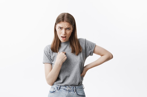 Gut aussehende junge kaukasische Studentin mit dunklem Haar und braunen Augen in grauem T-Shirt und Jeans, die mit dem Finger auf sich selbst zeigen, mit wütendem Ausdruck, wütend, wenn Mädchen in der Universität