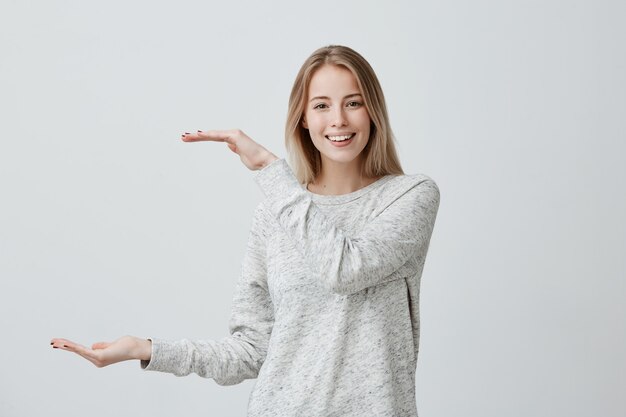 Gut aussehende schöne Blondine mit gefärbten Haaren Frau in Freizeitkleidung zeigt mit Händen Länge der Box. Ein zufriedenes weibliches Lächeln zeigt freudig die Größe von etwas Großem. Positive Emotionen und Gefühle