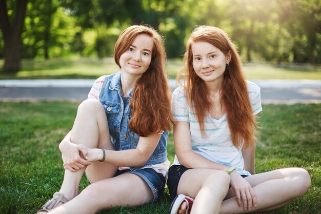 Gut aussehende zwei Frauen mit roten Haaren und Sommersprossen, die auf Gras nahe dem Universitätscampus sitzen und chillen