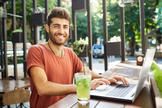 Gut aussehender, attraktiver bärtiger Mann, der breit in die Kamera lächelt, die lässig gekleidet am Holztisch sitzt, Limonade trinkt und Hochgeschwindigkeitsinternet auf Laptop-PC surft. Sommertag genießen.
