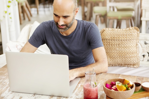 Kostenloses Foto gut aussehender erwachsener mann mit bart, der e-mail auf laptop überprüft, freie drahtlose internetverbindung im café nutzt und frühstückt.