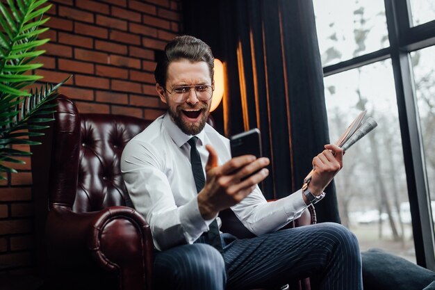 Gut aussehender Geschäftsmann mit offenen Emotionen, der sein Smartphone und seine Zeitung hält, während er auf einem Sessel sitzt.