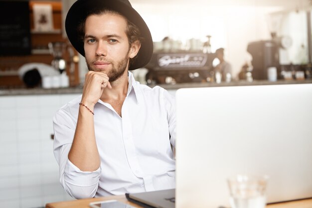 Gut aussehender junger selbständiger Mann mit Bart, der im Café vor modernem Laptop sitzt, seinen Ellbogen auf Tisch legt und ernsthaften fokussierten Blick hat, während er online am Notebook arbeitet