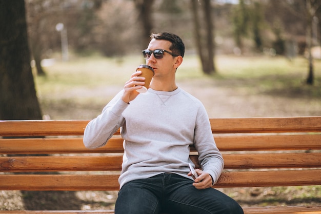 Gut aussehender Mann, der in trinkendem Kaffee des Parks sitzt und Telefon verwendet