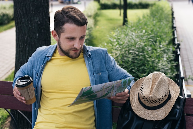 Kostenloses Foto gut aussehender mann, der wegwerfkaffeetasse beim ablesen der karte beim sitzen im park hält