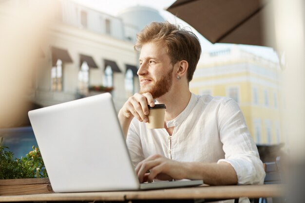 Gut aussehender Mann des erwachsenen Ingwers mit Laptop, der auf der Terrasse eines Restaurants oder Cafés sitzt