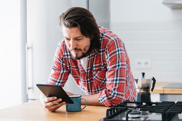 Gut aussehender Mann mit Tasse Kaffee, der Handy auf Küchenarbeitsplatte betrachtet