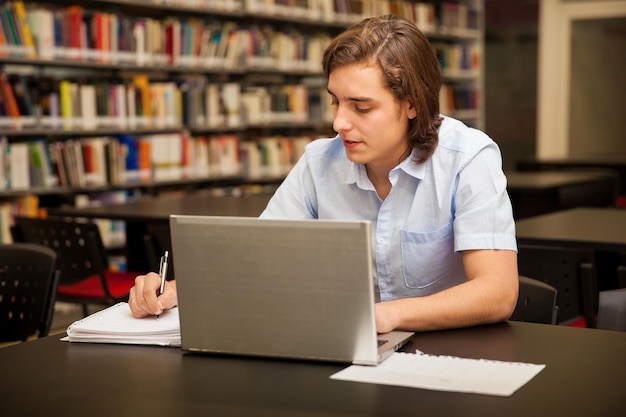 Gut aussehender Student, der sich Notizen macht und einen Laptop in der Bibliothek benutzt