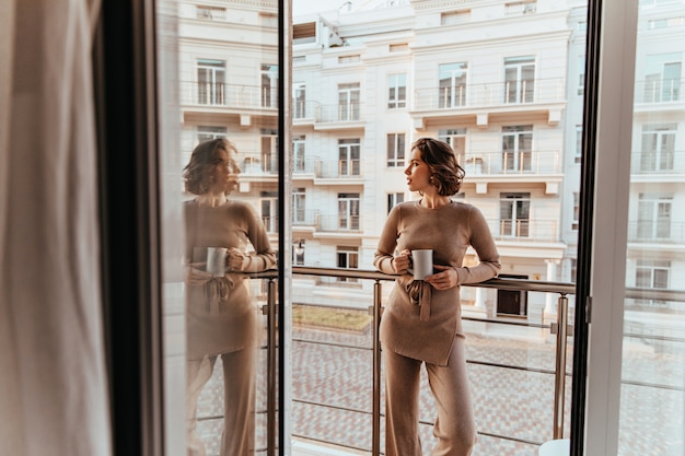 Gut gekleidete lockige Frau, die nahe großes Fenster mit Kaffee steht. Foto der herrlichen kaukasischen Dame, die Tee genießt und die Straße betrachtet.