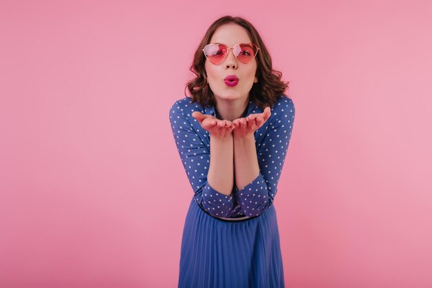 Gut gelauntes brünettes Mädchen, das einen Luftkuss sendet, während es auf pastellfarbenem Hintergrund posiert Schönes kaukasisches weibliches Model in blauer Kleidung, das positive Emotionen während des Fotoshootings ausdrückt