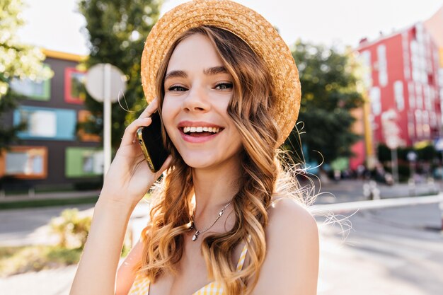 Gut gelauntes weißes Mädchen, das am Sommertag am Telefon spricht. Außenporträt der spektakulären Dame im Hut, der Smartphone auf Straße hält.