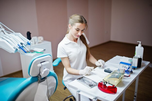 Gutaussehende Zahnärztin posiert mit einigen zahnärztlichen Instrumenten in der Hand in weißem Mantel in einem modernen, gut ausgestatteten Schrank