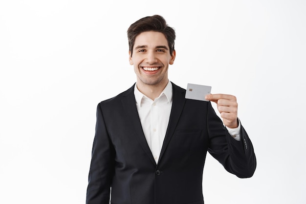 Gutaussehender ceo-manager eines unternehmens, der eine kreditkarte aus plastik zeigt und einen zufriedenen anzug trägt, der über weißem hintergrund steht
