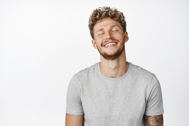 Gutaussehender lächelnder blonder Mann, der die Augen schließt und Tagträumen sieht, als er sich etwas vorstellt, das eine glückliche Erinnerung hat, die in einem grauen T-Shirt auf weißem Hintergrund steht