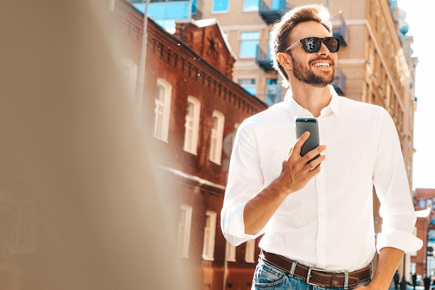 Gutaussehender, lächelnder, stylischer Hipster-Lambersexual-ModelModerner Mann in weißem Hemd Modemann posiert auf dem Straßenhintergrund im Freien bei Sonnenuntergang Mit Smartphone-Apps In Sonnenbrille