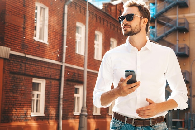 Gutaussehender, lächelnder, stylischer Hipster-Lambersexual-ModelModerner Mann in weißem Hemd Modemann posiert auf dem Straßenhintergrund mit Sonnenbrille Im Freien bei Sonnenuntergang Mit Smartphone-Apps