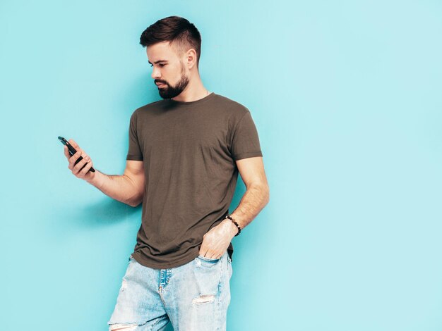 Gutaussehender, selbstbewusster, stilvoller Hipster-Lambersexual-ModelMann gekleidet in T-Shirt und Jeans Modemann isoliert auf blauer Wand im StudioHolding-Smartphone Blick auf Handy-Bildschirm Verwendung von Apps