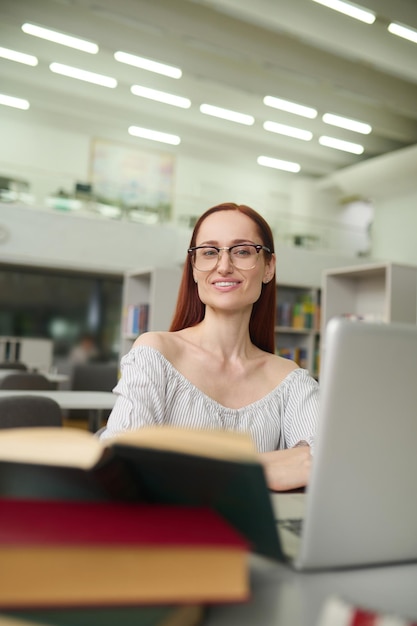 Gute Laune. Junge intelligente langhaarige Frau mit Brille in Bluse mit nackten Schultern, die am Tisch mit Laptop arbeitet und in einem beleuchteten Raum in die Kamera schaut