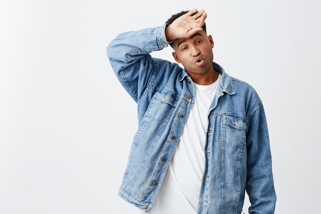 Gute ruhe nach langer arbeit. porträt des jungen schönen dunkelhäutigen amerikanischen mannes mit afro-frisur im weißen t-shirt und in der jeansjacke, die hand auf stirn hält und nach hartem tag müde ist.