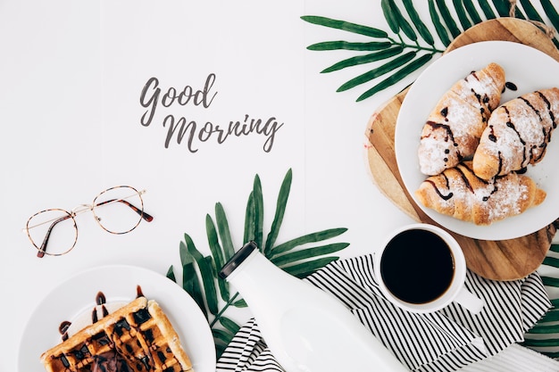 Guten Morgen Text mit Brille; frisch gebackenes Croissant; Waffeln; Flasche und Kaffeetasse auf weißem Schreibtisch