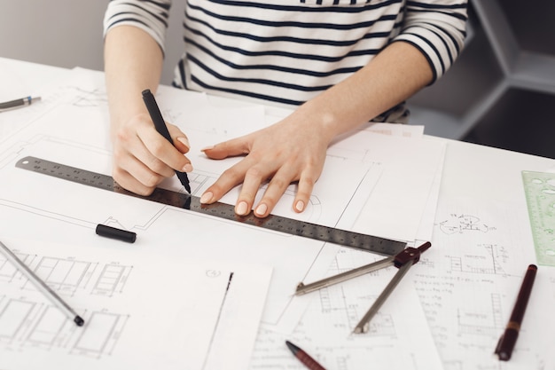 Hände der erfolgreichen jungen Architektin im gestreiften Hemd sitzen am weißen Tisch im Haus, machen Zeichnungen mit Stift und Lineal, Projekt ihres zukünftigen Zimmers machend.