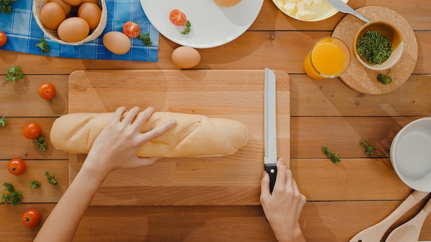 Hände der jungen asiatischen Köchin halten Messer, das Vollkornbrot auf Holzbrett schneidet