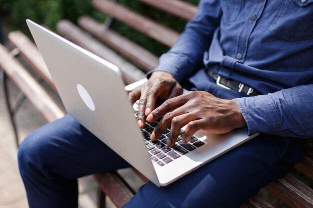 Hände des Afroamerikanermannes etwas auf dem Laptop schreibend, während er auf der Bank sitzt