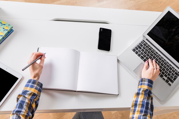 Hände des Frauenschreibens im Notizbuch und in der rührenden Tastatur des Laptops