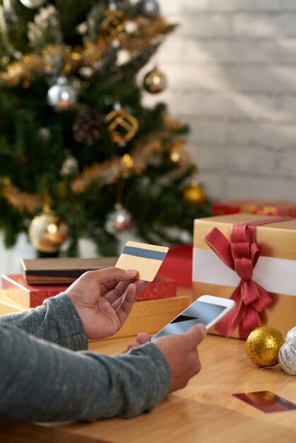 Hände des unerkennbaren Mannes Smartphone und Kreditkarte vor Weihnachtsbaum halten