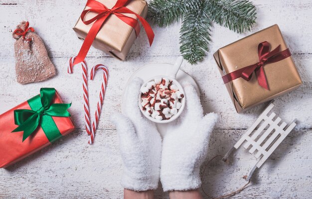 Hände, die eine Tasse mit Marshmallows und Weihnachtsgeschenke