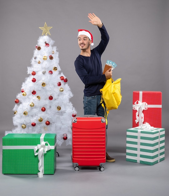 Hagelnder junger Mann mit Weihnachtsmütze und rotem Koffer, der seinen gelben Rucksack um verschiedene Geschenke prüft