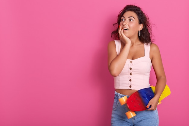 Halbe Länge der Aufnahme einer glücklichen, überraschten, lockigen Frau, die Zeit mit Freunden verbringt, bereit, gemeinsam Skateboard zu fahren, ein rosiges T-Shirt und eine Jeans trägt und mit offenen Augen posiert. Kopieren Sie Platz für Werbung.