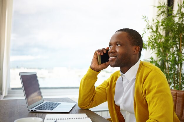 Halbe Profilaufnahme des schönen stilvollen afroamerikanischen Designers, der auf Handy mit dem Kunden spricht, Details und Ideen des Hausinnenprojekts bespricht, inspirierten Blick, am Fenster sitzend