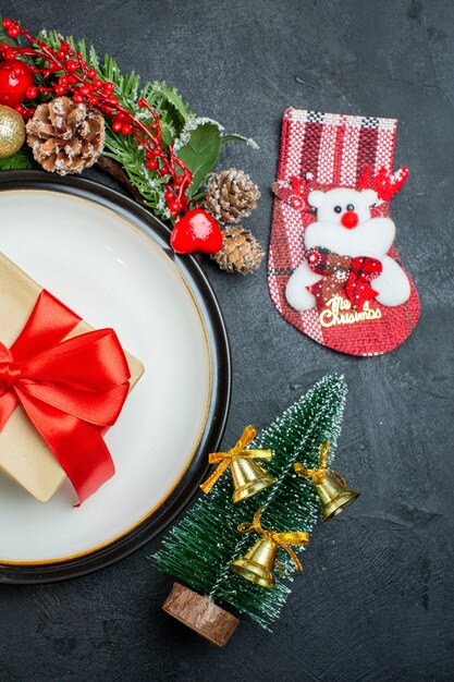 Halbe Schussansicht der Geschenkbox auf dem Teller der Weihnachtsbaum-Tannenzweige Nadelbaumkegel-Weihnachtsmannhut gefallener Glasbecher auf dunklem Hintergrund