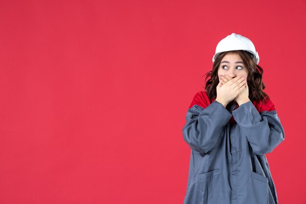 Halber Körperschuss einer schockierten neugierigen Baumeisterin in Uniform mit Schutzhelm auf isoliertem rotem Hintergrund