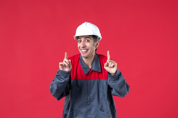 Halber Körperschuss eines glücklichen jungen Baumeisters in Uniform mit Schutzhelm, der auf isolierte rote Wand zeigt