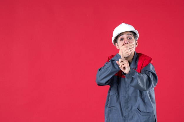 Halber Körperschuss eines verängstigten jungen Architekten in Uniform mit Schutzhelm und nach oben auf isolierte rote Wand zeigend