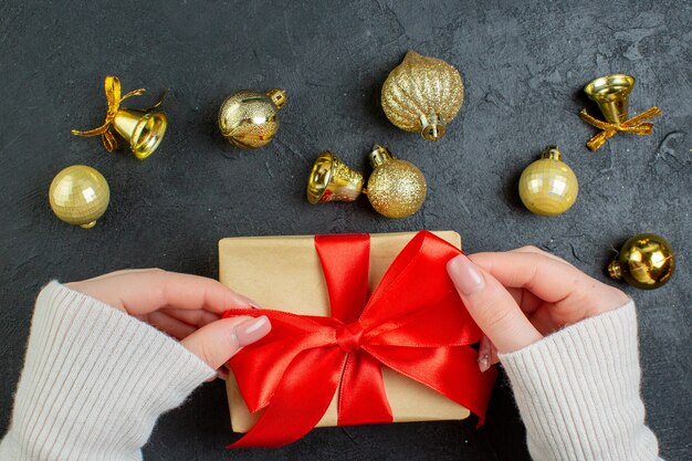 Halber Schuss der Hand, die eine der Geschenkboxen mit rotem Band und Dekorationszubehör auf dunklem Hintergrund hält