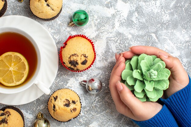 Halber Schuss einer Tasse schwarzen Tee mit Zitrone unter frisch gebackenen köstlichen kleinen Cupcakes und Dekorationszubehör und Hand, die Nadelbaumkegel auf Eisoberfläche hält