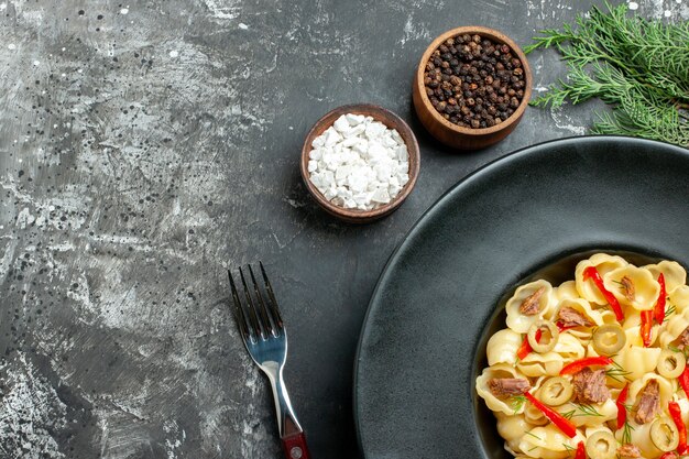 Halber Schuss köstlicher Conchiglie mit Gemüse und Grün auf einem Teller und Messer und verschiedenen Gewürzen auf grauem Hintergrund