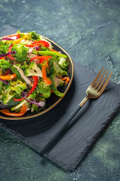 Halber schuss köstlicher veganer salat mit frischen zutaten in einem teller und einer gabel auf schwarzem schneidebrett auf blauem hintergrund