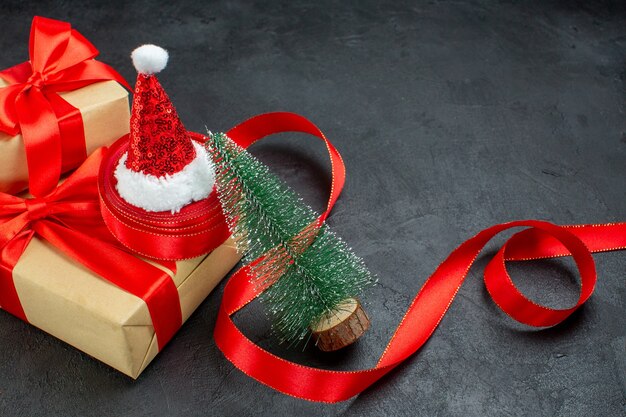 Halber Schuss von schönen Geschenken mit rotem Band und Weihnachtsmannhut-Weihnachtsbaum auf dunklem Tisch