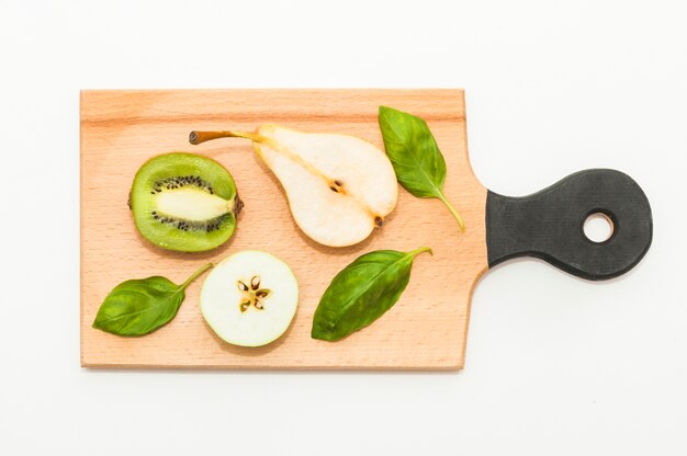 Halbierte Kiwi; Birnen; Apfel und Basilikum auf Schneidebrett vor weißem Hintergrund