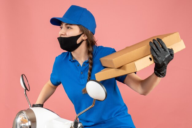 Halbkörperaufnahme einer jungen neugierigen Kurierin mit medizinischer Maske und Handschuhen, die auf einem Roller sitzt und Kisten auf pastellfarbenem Pfirsichhintergrund hält holding