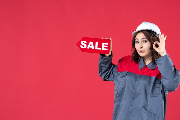 Halbkörperaufnahme einer lächelnden Arbeiterin in Uniform, die einen Schutzhelm trägt und ein Verkaufssymbol zeigt, das eine Brillengeste auf isoliertem rotem Hintergrund macht